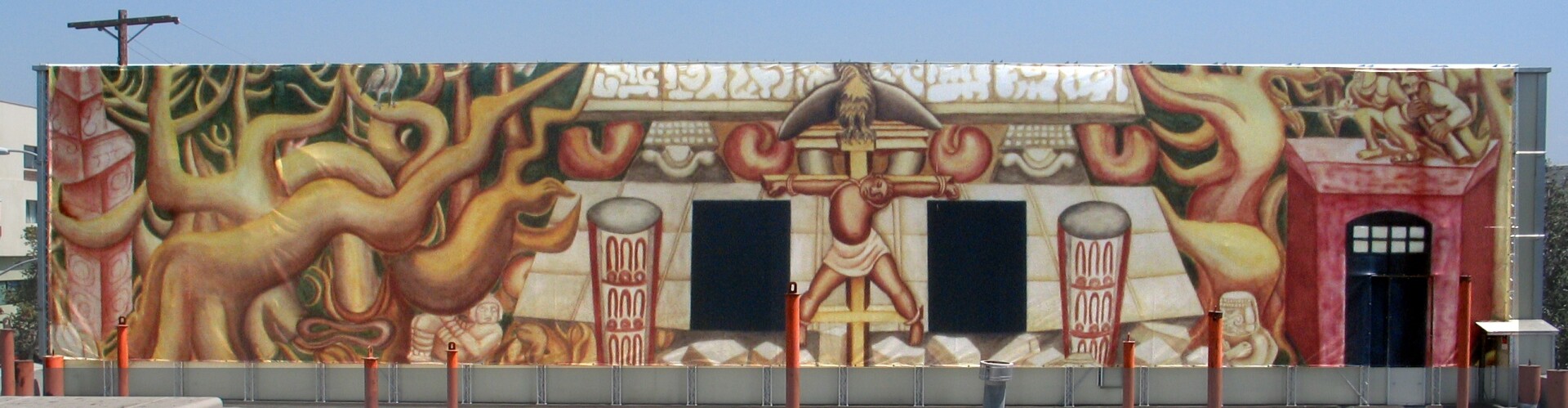 A photo of a mural depicting a a crucified Indigenous worker. Above, a screaming eagle is perched on top of the cross and crouching snipers aim at the bird. The mural is painted in shades of reds, yellows, creams and a hint of forest green.