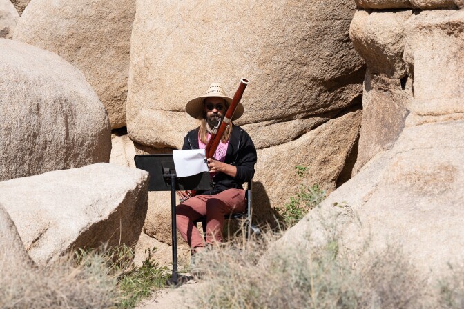 Archie Carey wearing a wide-brimmed straw hat and sunglasses sits on a chair and plays the bassoon, a long wind instrument that is held upright. A music stand is propped up in front of him as he looks on at the sheet music. Archie is playing the bassoon amidst large, rock desert formations. Desert flora grows in small nooks and crannies within the formation.