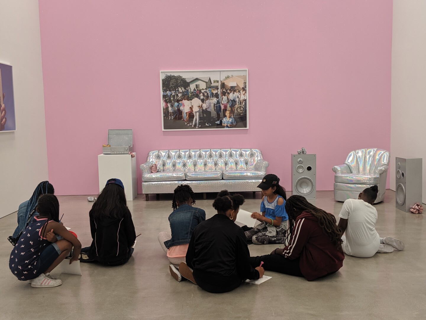 Children sit on the floor in front of a pink wall with artwork hung on it. Below the framed artwork are holographic and metal furniture.
