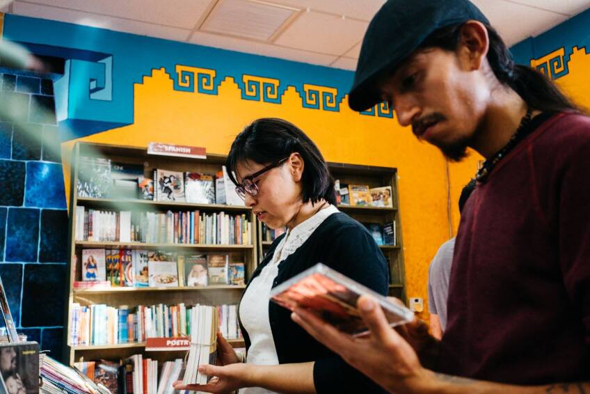 Two people standing side by side inspect a book and CD, respectively. Behind them is a bookcase pushed up against a vibrant yellow wall with teal blue detailing at the top. 