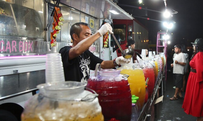 Fruit juices in jugs are lined up on a table in front of a food truck. A man in a black t-shirt uses a ladle to scoop juice into a small sample cup for a customer.