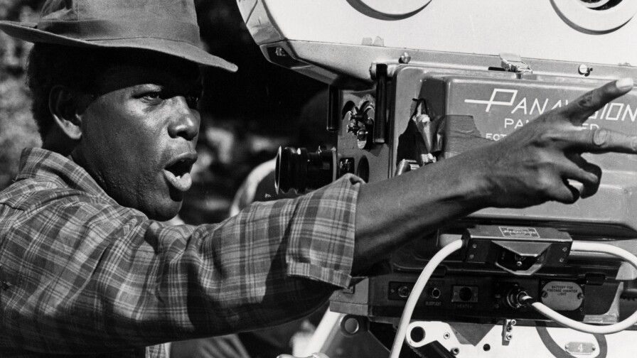 An old black and white photo of Sidney Poitier directing behind a large film camera. He's wearing a brimmed hat that partially shades his face and a plaid collared shirt. Poitier looks directly in front of him and points two fingers at something out of frame.