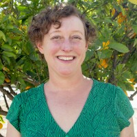 A headshot of writer Elaine Lewinnek. They have short, curly light brown hair. They're wearing a patterned green and blue top and they're standing in front of an orange tree.