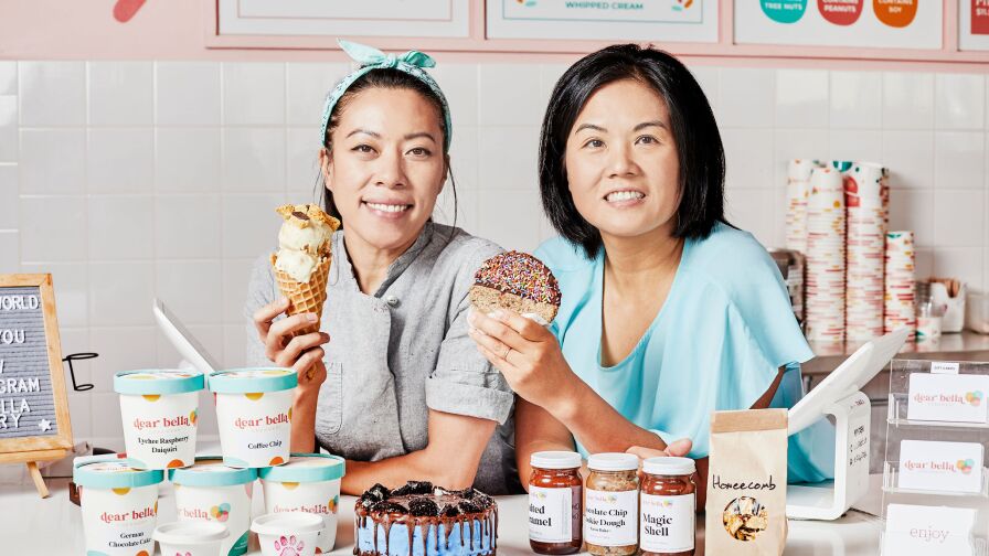 Belinda Wei and Alice Cherng lean over a counter topped with pints of ice cream, jars of ice cream toppings and a cake. Belinda, on the left, is holding an ice cream cone topped with two scoops of ice cream. Alice, right, holds an ice cream sandwich dipped in chocolate and sprinkles. The set and the two women are dressed in pastels.