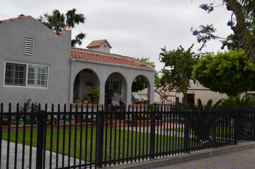 A gray stucco house with an arched patio entrance and two arched windows. The house has red roof tiling and plants all along the patio. A well-trimmed green lawn and a black iron-wrought fence surround the house.