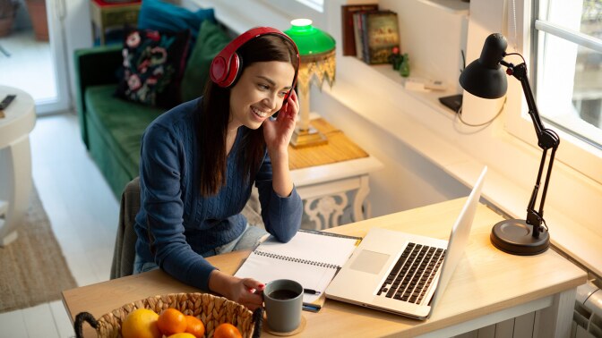 A young female teacher wearing red headphones works at a desk with a laptop on it.
