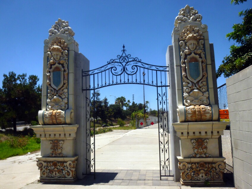 Two Beaux Arts-style pillars stand at the entrance to a concrete paved walkway into a park. Between the two pillars are iron fencing with ornate and intricate details. The pillars are carved with ornate shapes and colored in various oranges, golds and blues.