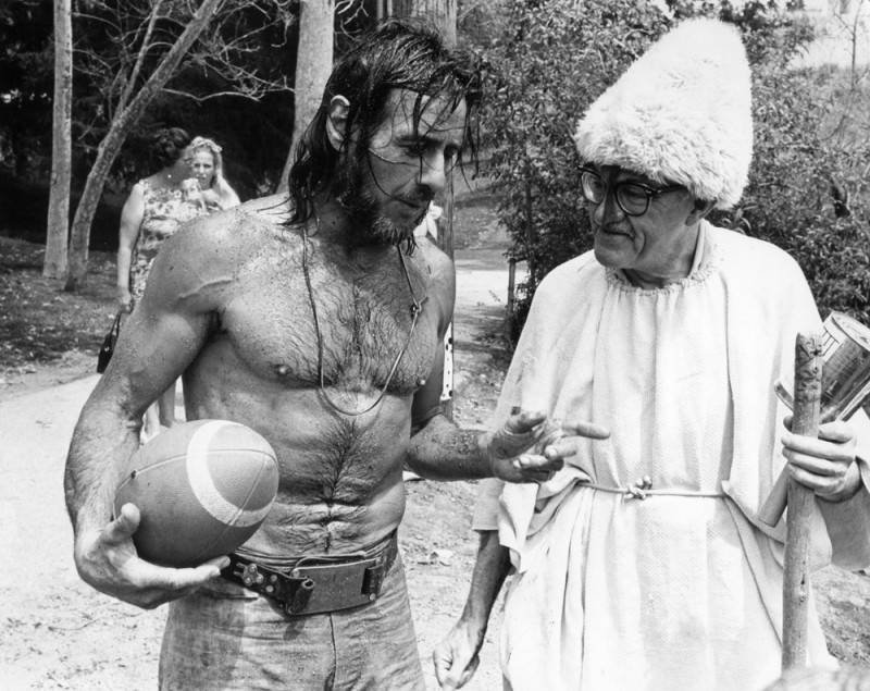 A black and white photo of Gypsy Boots, a football under his arm, conferring with Everett Goodelle, dressed in a Buddhist toga, and toting a prayer wheel. Mud and water cling to Gypsy's skin after a steam bath. The two are walking through a natural landscape.