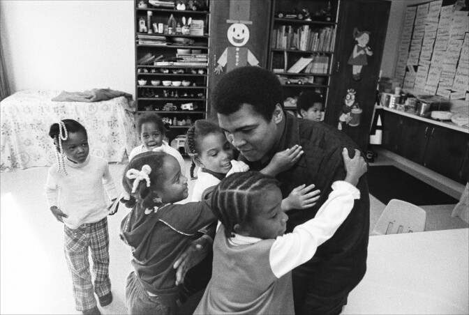 A Black man is hugged by other children in a schoolroom space.