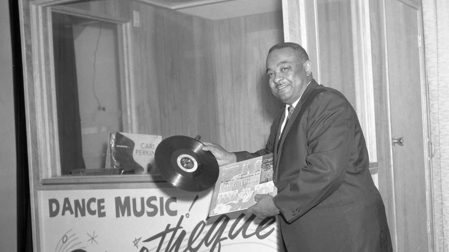 An African American man holds a record while leaning on a music booth.