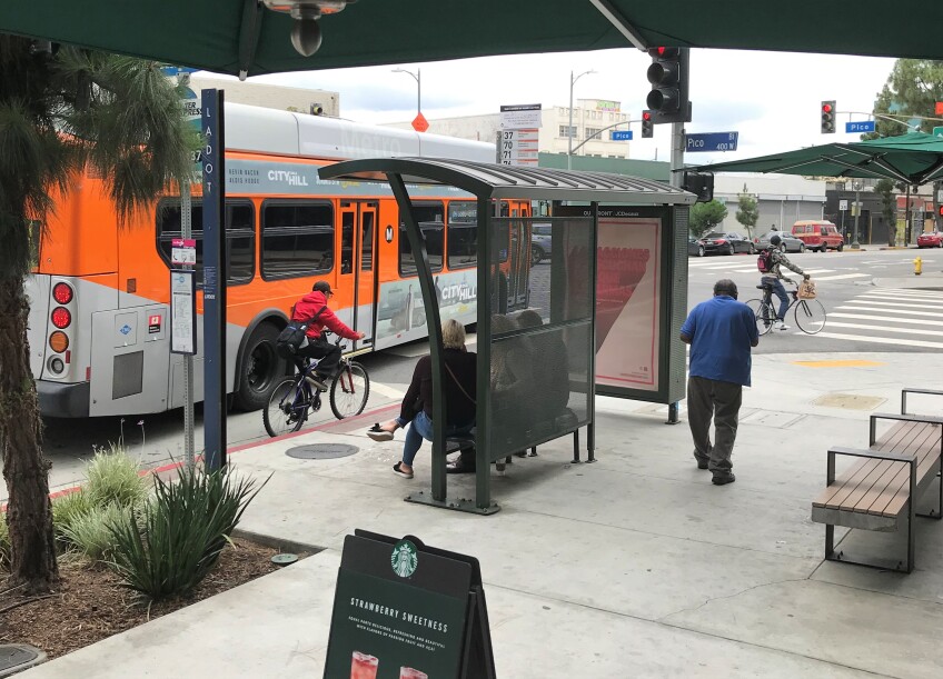 An L.A. Metro bus pulls up to a bus stop with a shelter, while commuters sit on a bench next to advertising.