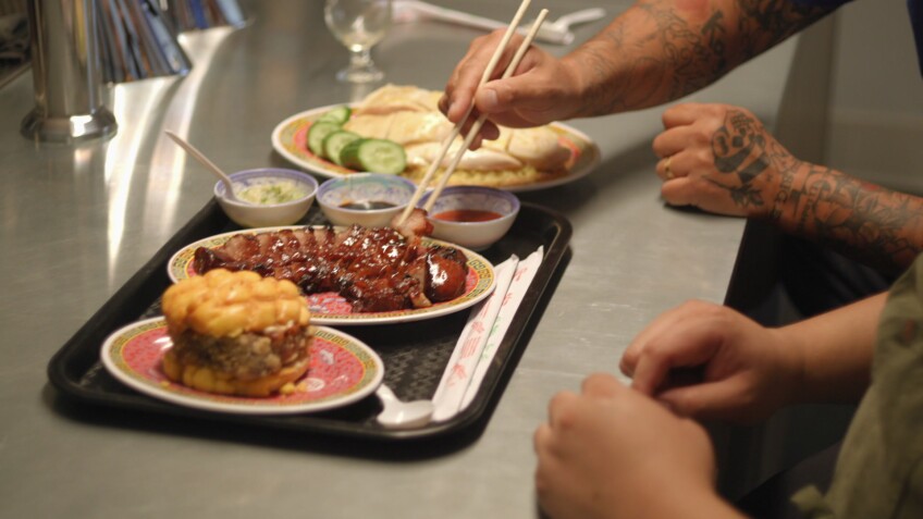 Cantonese comfort food served on dishes over a plastic tray and stainless steel counter. From left to right: a small plate topped with breaded pork chop sandwiched between yellow bolo bao buns; red roasted char siu pork sliced into pieces; three small sauce bowls lined up next to one another; and a large plate of poached Hainanese chicken over a bed of golden yellow rice.