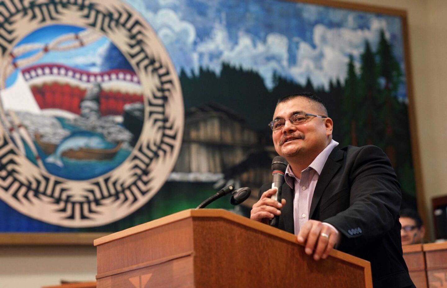A man in a black suit jacket and glasses speaks at a podium with a Yurok seal in the background.