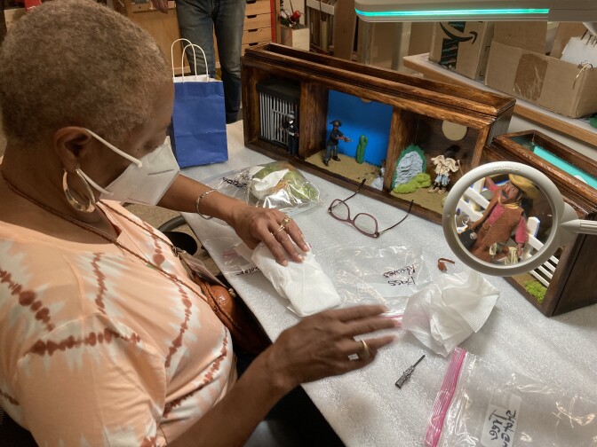 Karen Collins is wearing a pink tie-dye shirt, gold earrings, and a white medical mask. An incomplete three-panel diorama sits on the table in front of her. Other materials for the diorama are sprawled out on the table such as fake foliage in a plastic bag. Her eyeglasses sit on the table as well. A magnifying glass magnifies a small clay figure in the diorama.