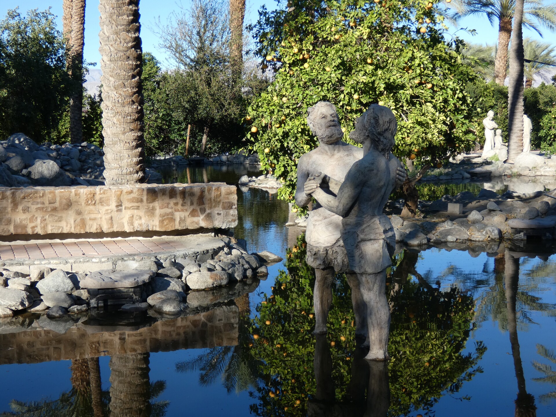 Two statues of people stand side by side, hand-in-hand in a pool of still water. The pool is surrounded by date palm trees, which are reflected in the still water. 