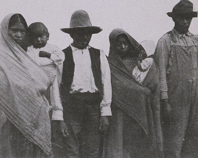 Two Mexican families in a posed black and white picture. From left to right is a woman holding a small child, a man wearing a tall hat, another woman holding a baby and a taller man in overalls and a short hat.