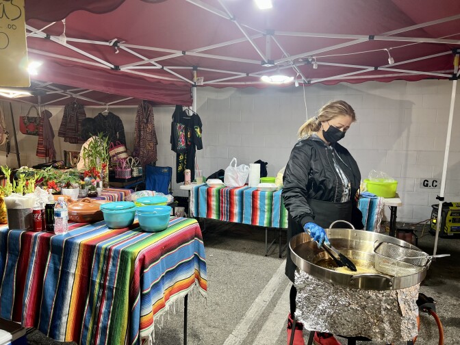 Minerva Orsorno stands over a big pot of hot oil. With a pair of tongs, she turns a quesadilla over in the bubbling oil. Behind her are two tables covered with multicolor tablecloths. A red tent stretches above her.
