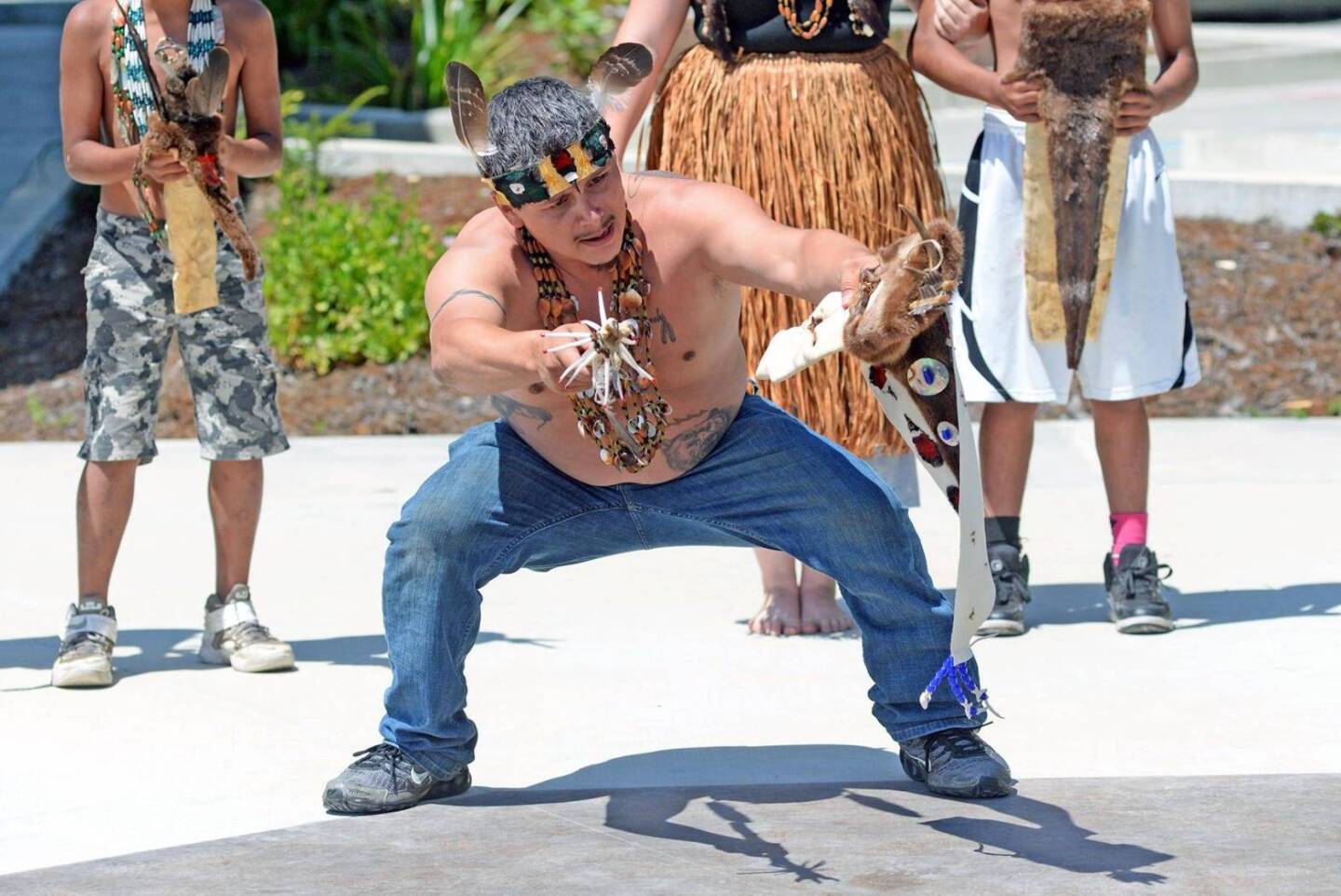 A shirtless man in jeans and sneakers wears Indigenous Yurok decorations like a necklace and headband as he performs a Brush Dance demonstration outdoors.