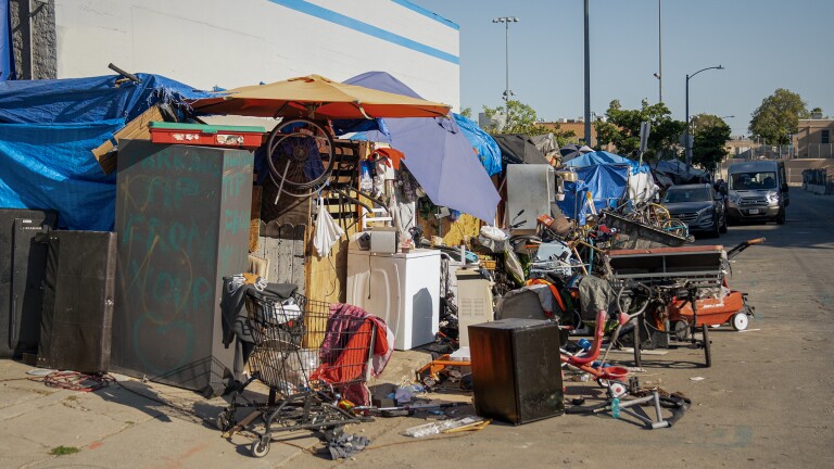 A homeless encampment consisting of umbrellas, tarps, a shopping cart, a washing machine and other debris spills out from the sidewalk and into the street, in front of parked cars.