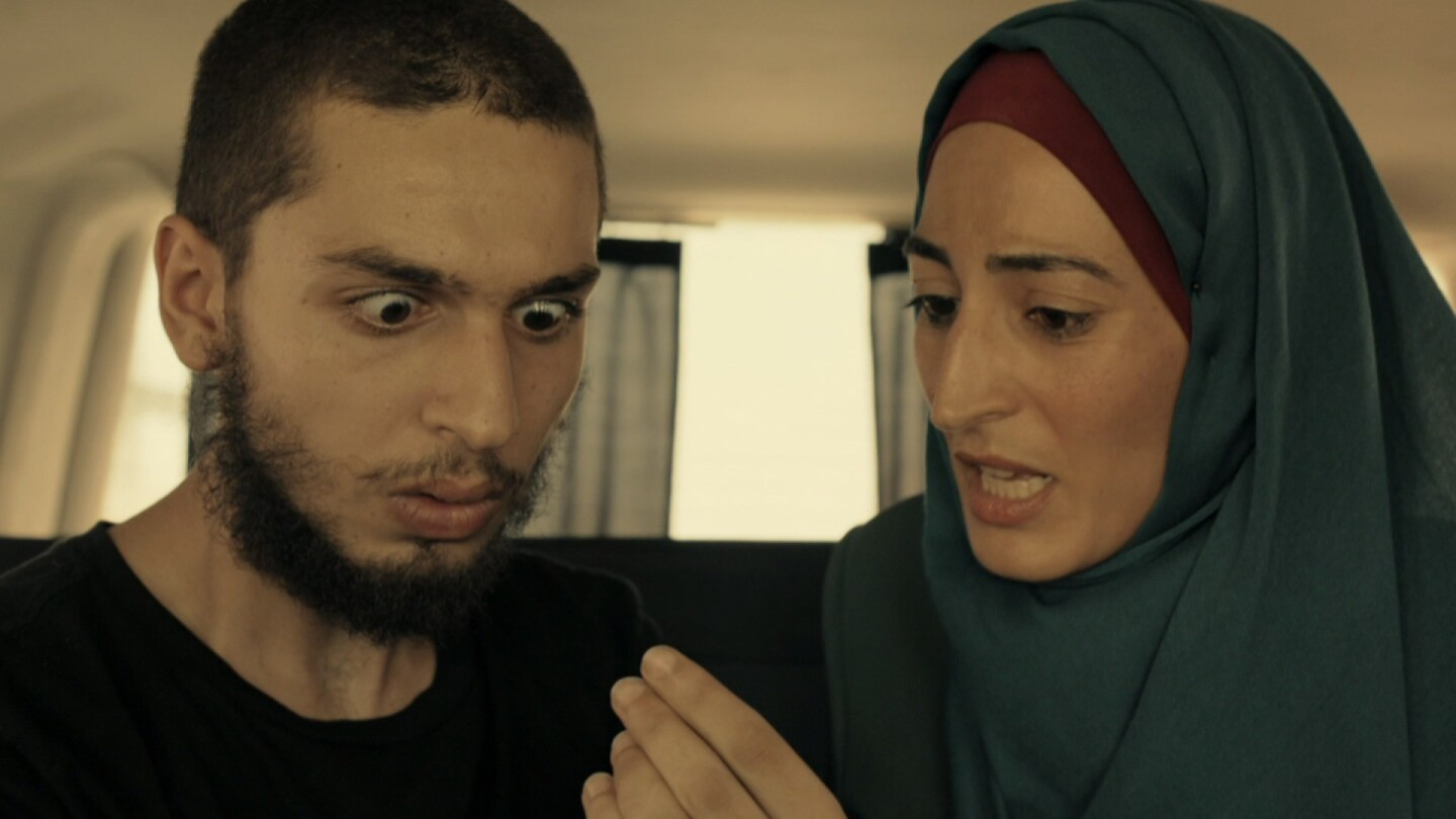 Man with his eyes widened at something a woman wearing a hijab is holding. | From "Fauda: S2 E6."