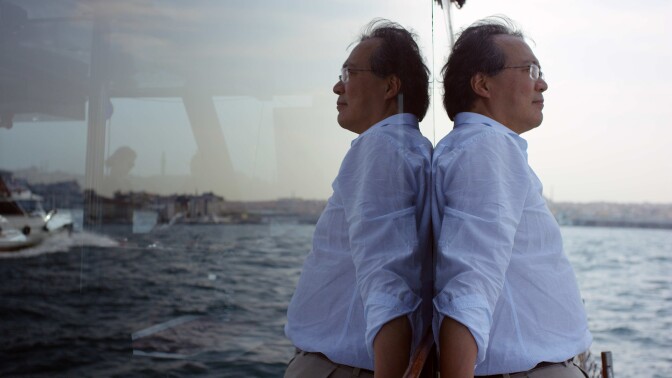 Yo-Yo Ma leans against a wall of glass showing a reflection of the sea. | From "The Music of Strangers"