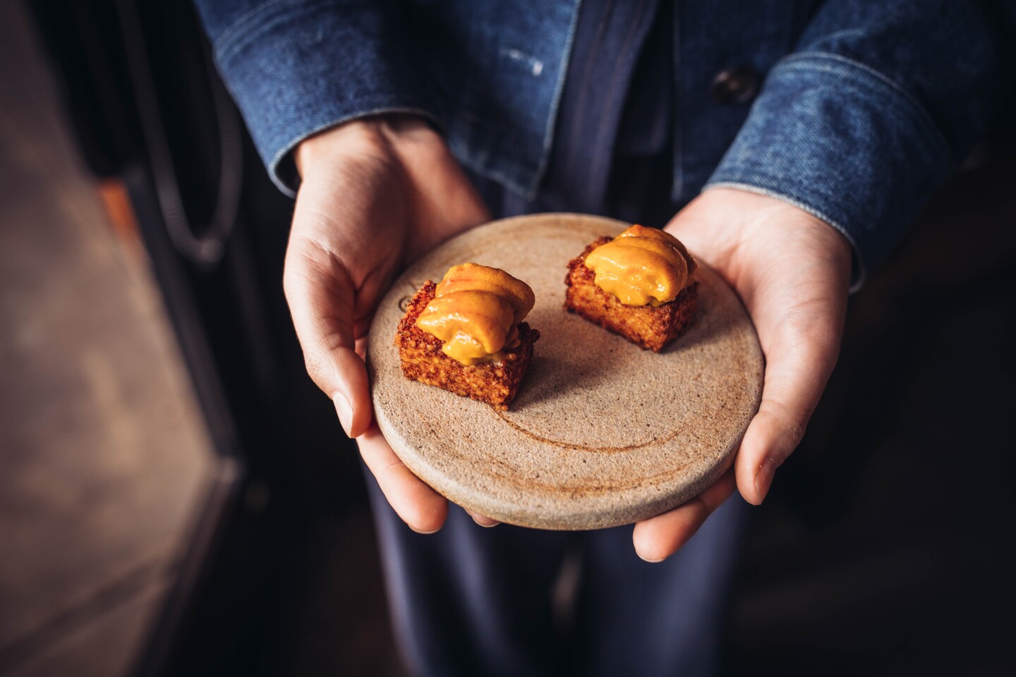 Two bite-sized food sit on a small, smooth ceramic plate. An orange, creamy slab of umi sits on top of a cube-shaped deep-fried tapioca.