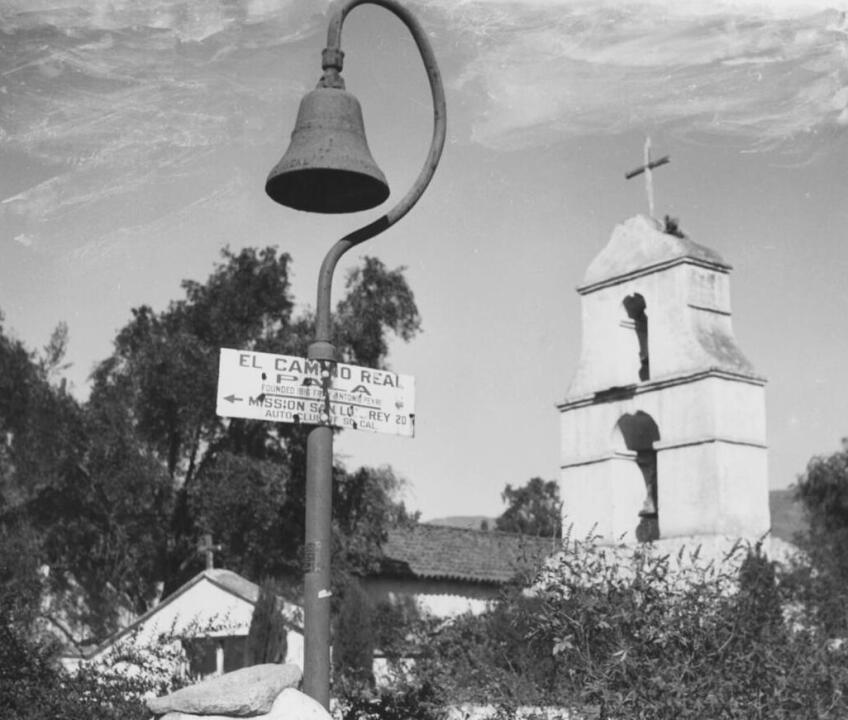 Photograph of Mission Asistencia at Pala, with Camino Real Bell, circa 1900. A bell that hangs from a curved pole stands to the left of center witha sign attached that reads "El Camino Real Pala Founded 1816 Fr..y Antonion Peyri Mission SanLu..rey 20 Auto Club Of So Cal." A bell tower can be seen at right with two bells and a cross on top. A small building with a cross is visible at left while trees can be seen all around.