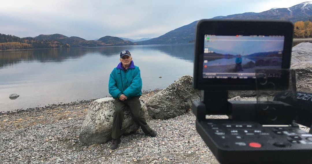 An elderly man in a teal windbreaker jacket sits on a rock. Behind him is a scenic landscape consisting of a serene lake and rolling mountains and hills dotted with trees of different shades — oranges, yellows, greens.