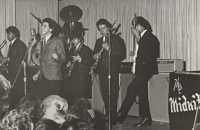 A black and white photo of Thee Midniters performing on a stage. A man stands in the middle, singing into a microphone on a mic stand. Three men stand behind him, two with an electric guitar and one with a bass. A saxophonist is off in the distance and a drummer plays a drum set on a raised platform. At the performers' feet, you can see the back of women's heads, looking up at the performance.