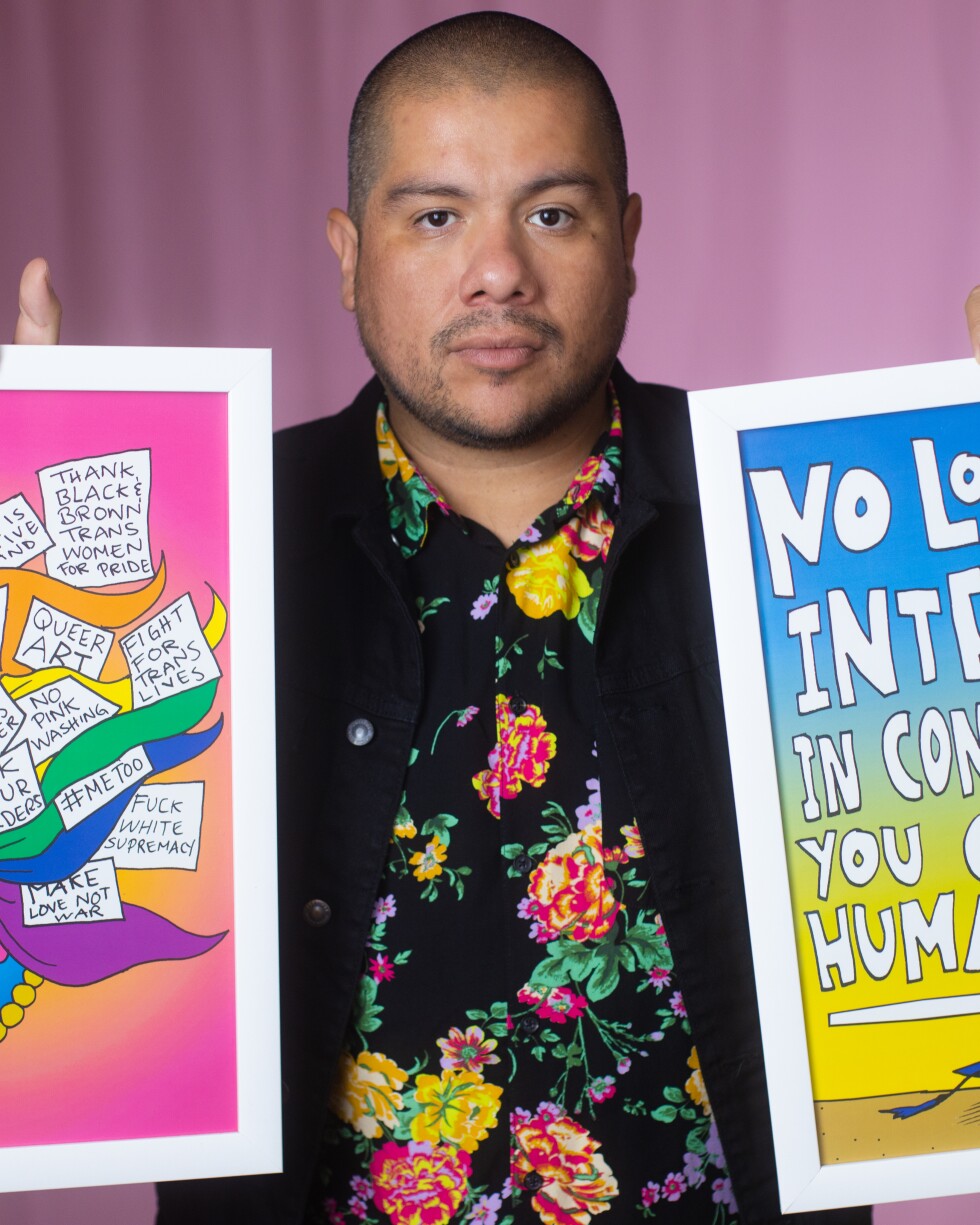 Julio Salgado is wearing a floral print shirt and a black jacket while holding up two pieces of his art on each hand. The artwork on his left features the side profile of a woman with multicolored hair and statements like, "Black Lives Matter," "#MeToo," "Make Love Not War," and "Thank Black and Brown Trans Women for Pride." The artwork on the right reads, "No Longer Interested in Convincing You of My Humanity," with a graduation cap at the bottom. Salgado is standing in front of a pink background.