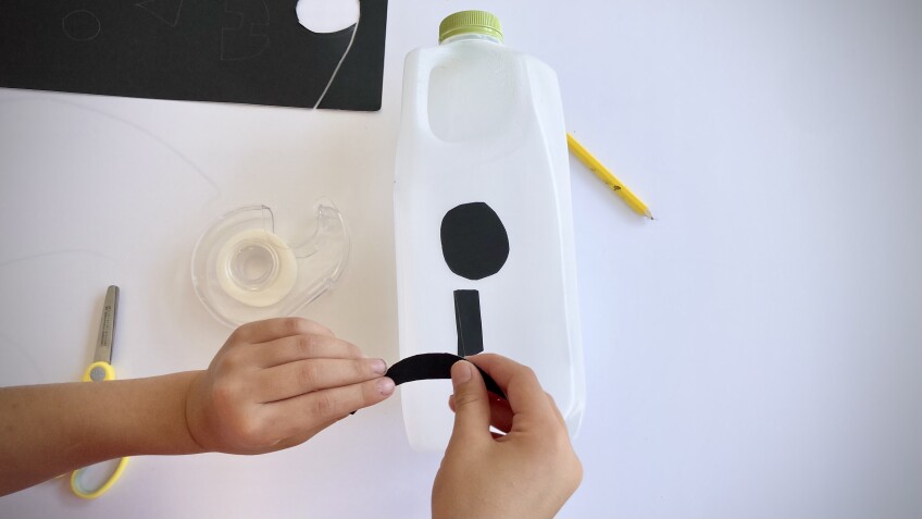 a child tapes on black construction paper shapes on an empty milk jug to form a jack o' lantern face