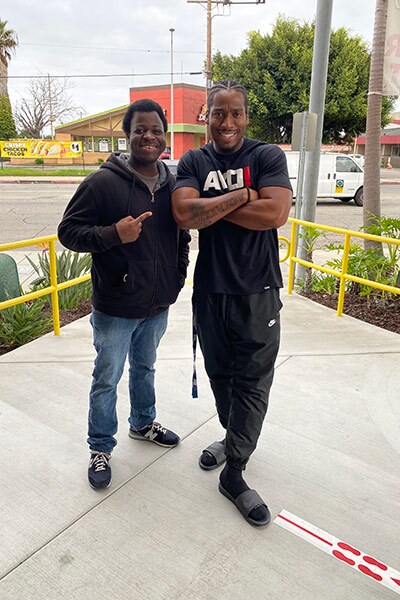 Two young people smile while posing for a photo on a sidewalk surrounded by a yellow rail. 