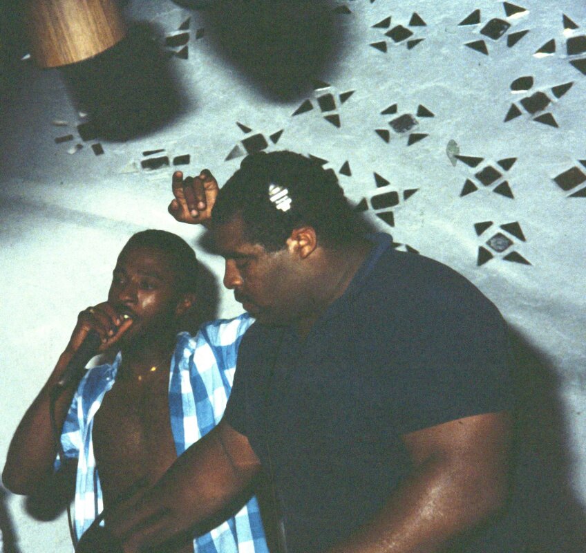 Two young African American men in a dance party. One holds the mic and the other spinning discs.