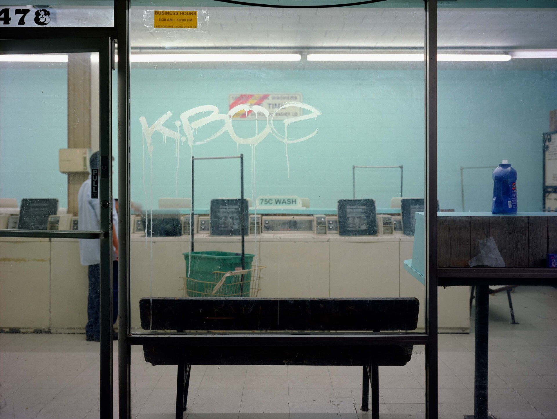 A laundromat taken from the outside. The floor to ceiling windows provide a complete view of the laundromat with a light blue wall. A bench is pushed up against one of the windows. The window has graffiti on it, reading "KBOE." A man is seen standing in front of one of the washing machines.