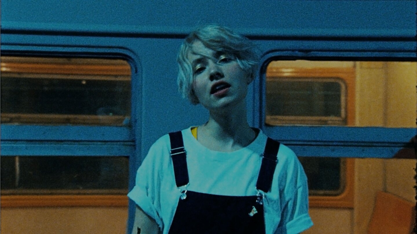 Still from NV's "Kata": A woman with short hair, a white T-shirt and overalls sings in front of a train car.