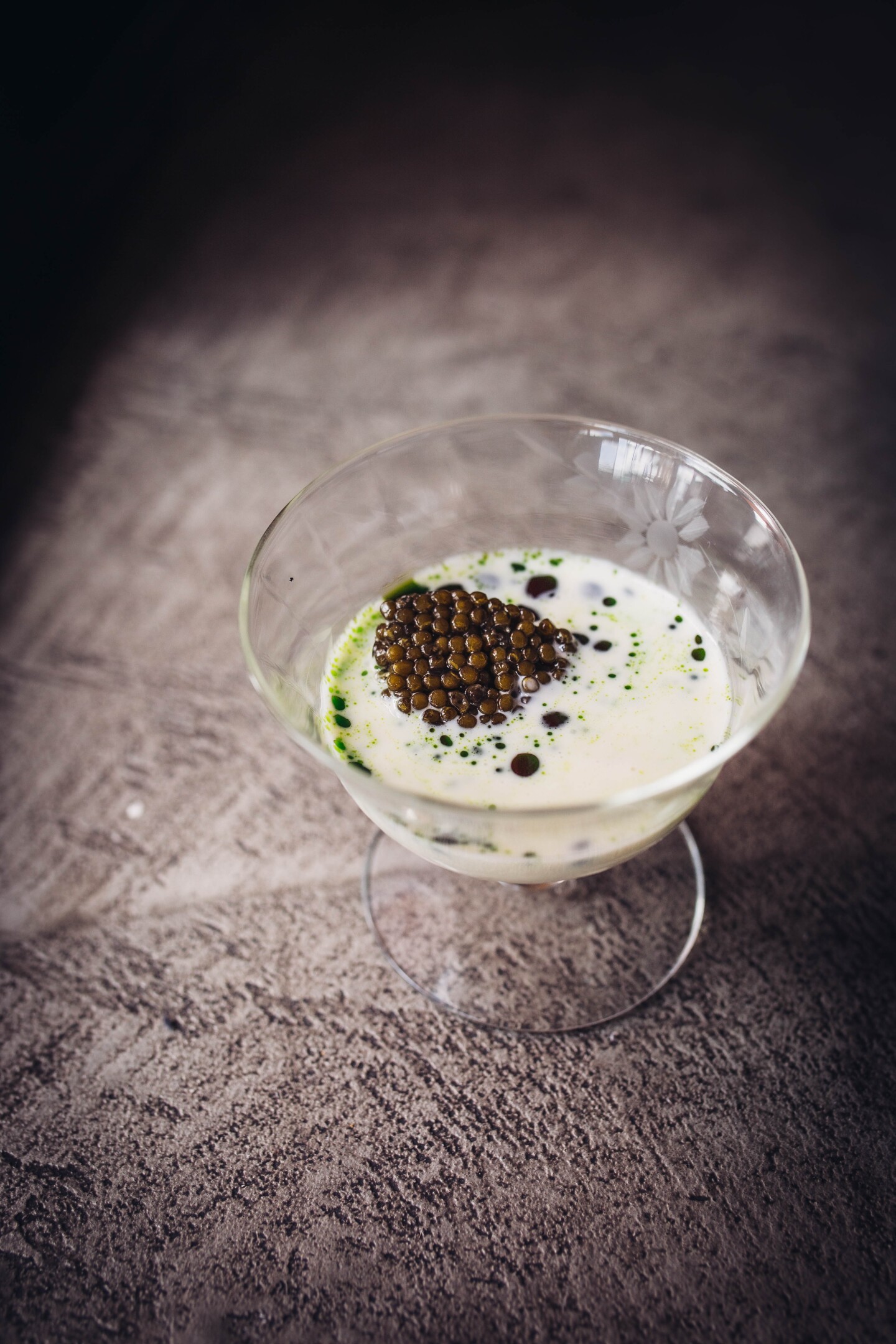 A small, cone-shaped glass holds a white chowder with spots of dark green oil sitting on top. A brown-colored caviar tops the dish.