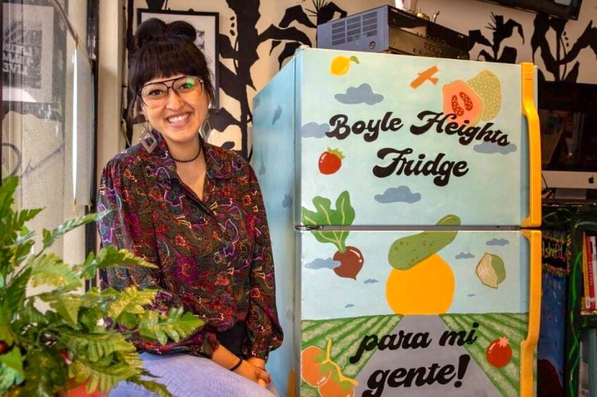 Deysi Serrano sits next to a bright refrigerator. A colorful, vibrant scene is painted on the fridge — a long road cuts between rows of green fields. The road leads to a bright orange sun as it sets on the horizon. The sun is set against a bright blue sky with dark blue clouds spread out throughout the scene. Colorful fruits and vegetables like pomegranates, radish, strawberries and lemons fall from the sky and a bird flies overhead.