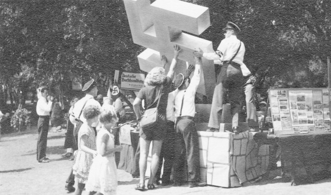 An archival black and white photo depicts people raising a large Swastika sculpture on a pedestal at a park.
