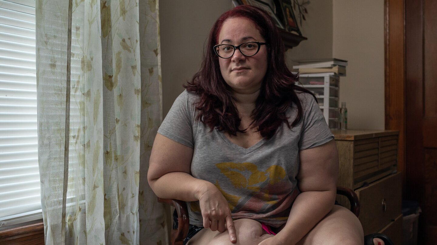 Monica sits at a chair next to a window lighting her face. She's sitting with one leg folded on the chair.