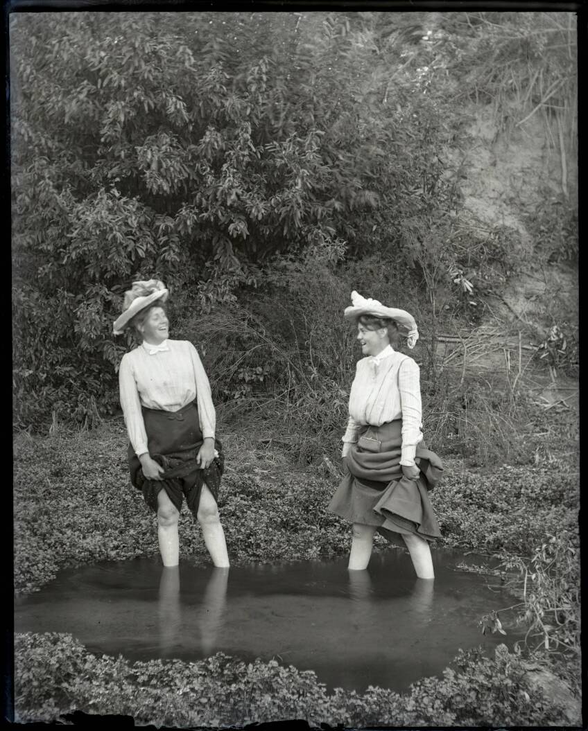 Two women in Victorian fashion stand up to their ankles in the murky water of Toluca Lake with their skirts hiked up above their knees. They both have big smiles on their faces.