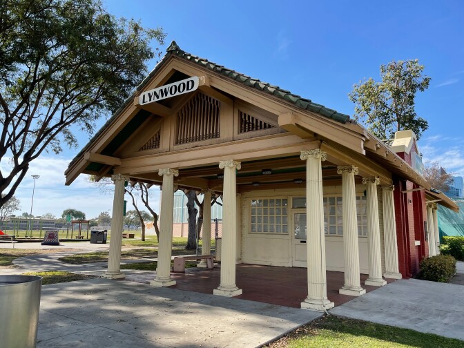 Lynwood Station, a small former train depot with ornate ivory columns and a small sign across the roof that reads, "Lynwood." The station stands amidst a public park and against a bright blue sky.