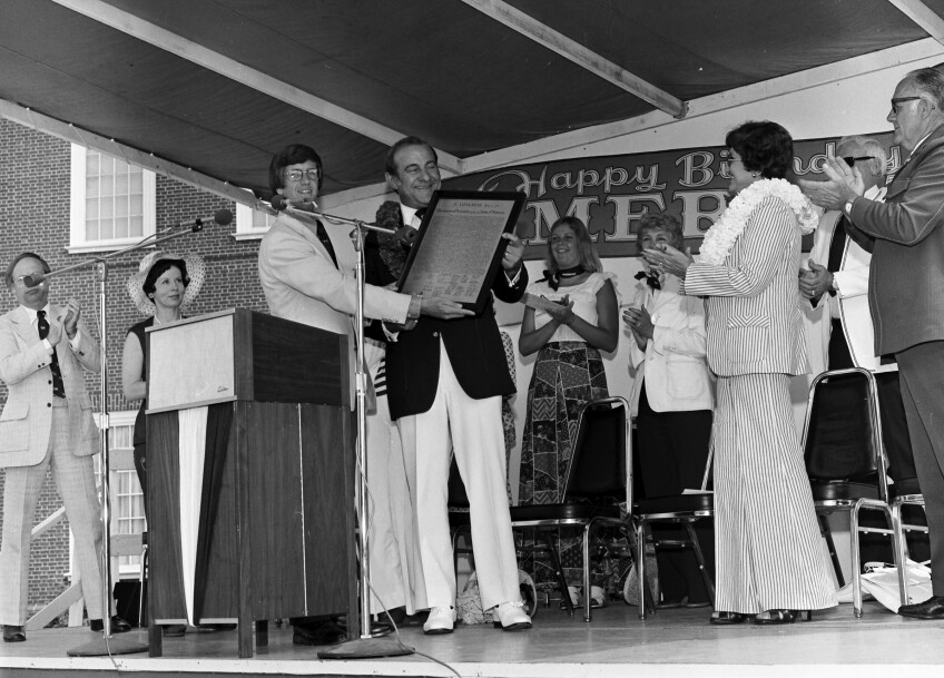 A man is presented with a framed document on a stage as onlookers applaud.