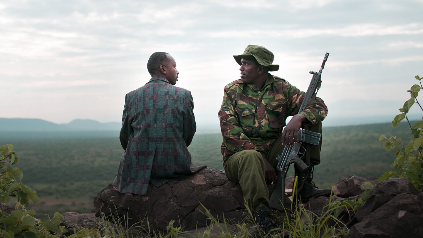 A man in civilian clothes looks at another man wearing an army uniform and resting a rifle in his arm. | "When Lambs Become Lions"