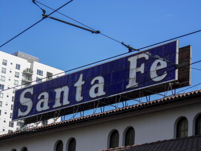 An old, blue and white sign that reads "Santa Fe" is mounted on top of a white building with red-tiled roofs. The letters that spell, "Santa Fe" are white and in a serif font over a blue background.