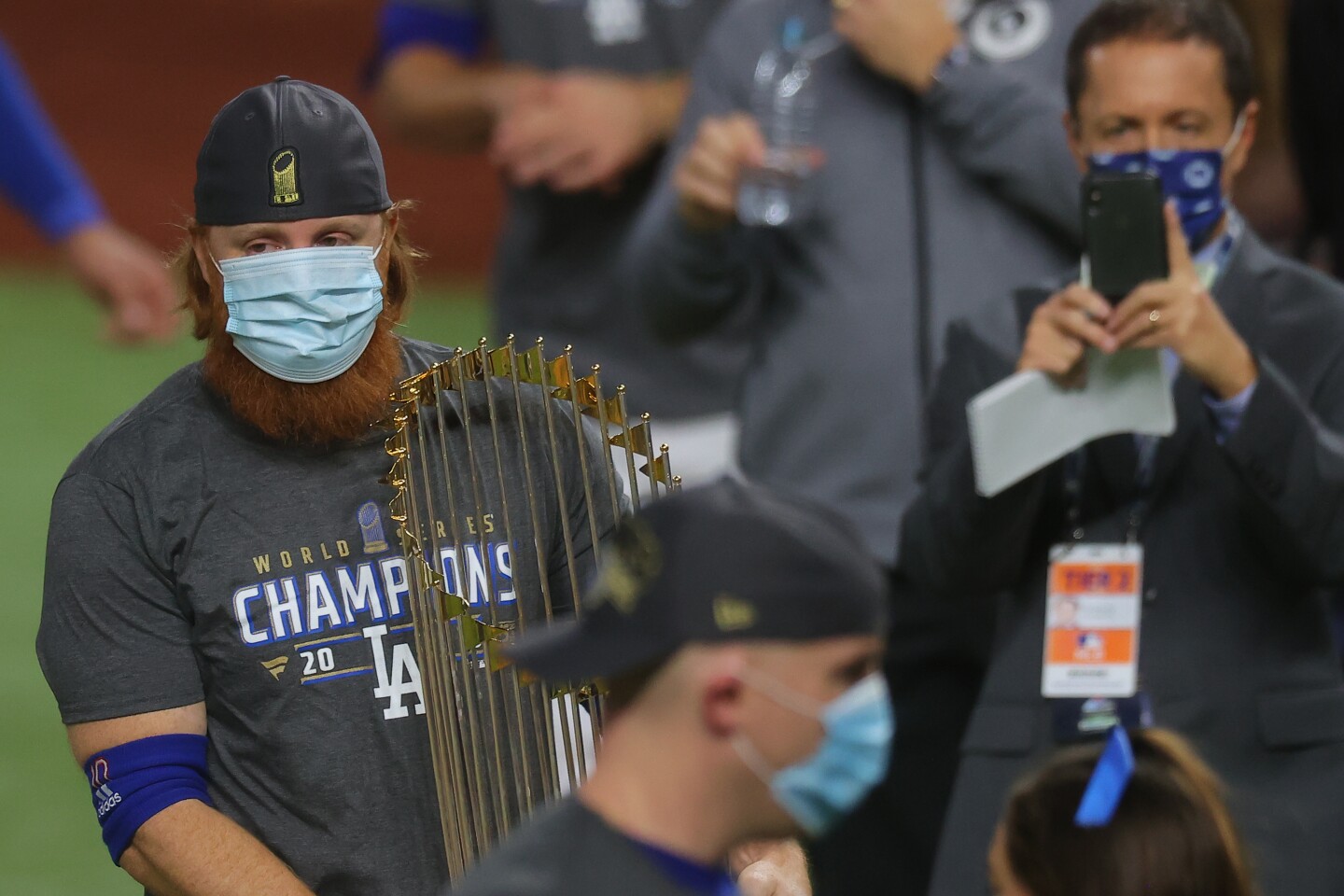 A red-headed and bearded man wearing a face mask holds a golden trophy on a baseball field.