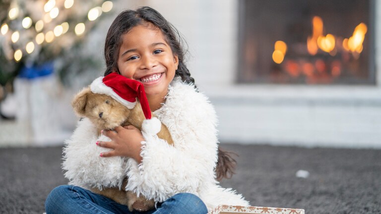 A small child sits in front of a fireplace and a tree with twinkly lights. She is holding a teddy bear and smiling. iStock