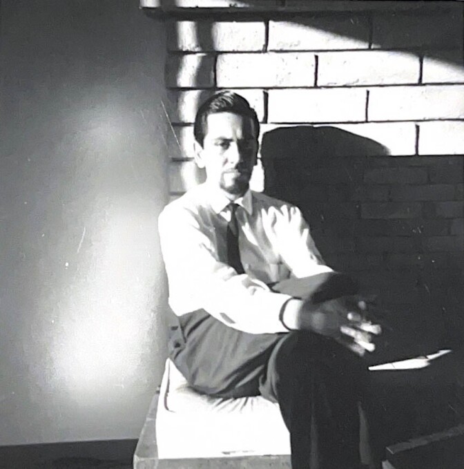 An archival black and white photo of J. Sergio O’Cadiz Moctezuma sitting on the mantle of a fireplace. His leg is crossed over the other and his hands are folded over his knee. He's wearing black slacks and a black tie and looks directly at the camera.