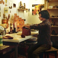 Lizette Hernández is seated in front of a table, sculpting a container out of clay, using her hands. She is wearing a button-up plaid shirt and her hair is cut short in a bob. She's surrounded by lit candle sticks on the table and art materials and projects in various stages of completion. 