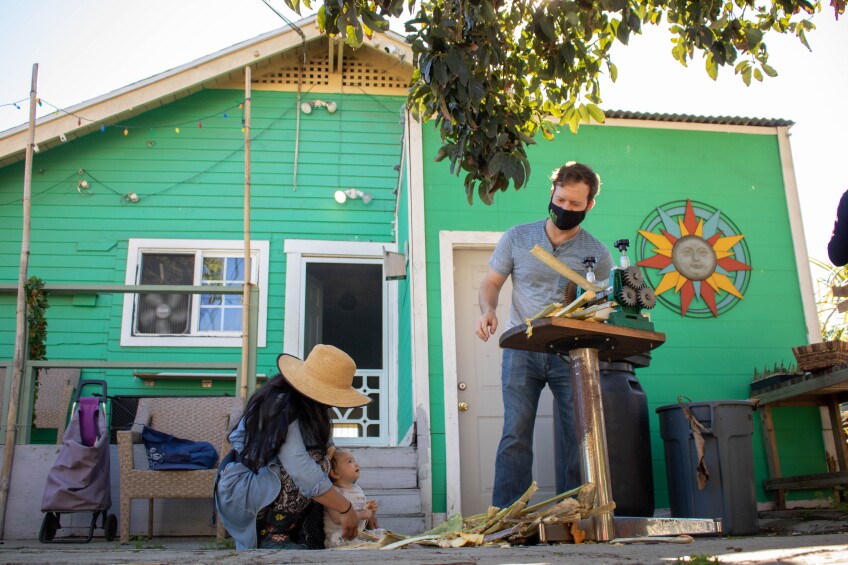 A woman squats over a baby sitting next to a pile of pressed sugar cane waste. The baby looks up at the sugar cane press on the table above being cranked by a young man. Behind the scene is a teal house.