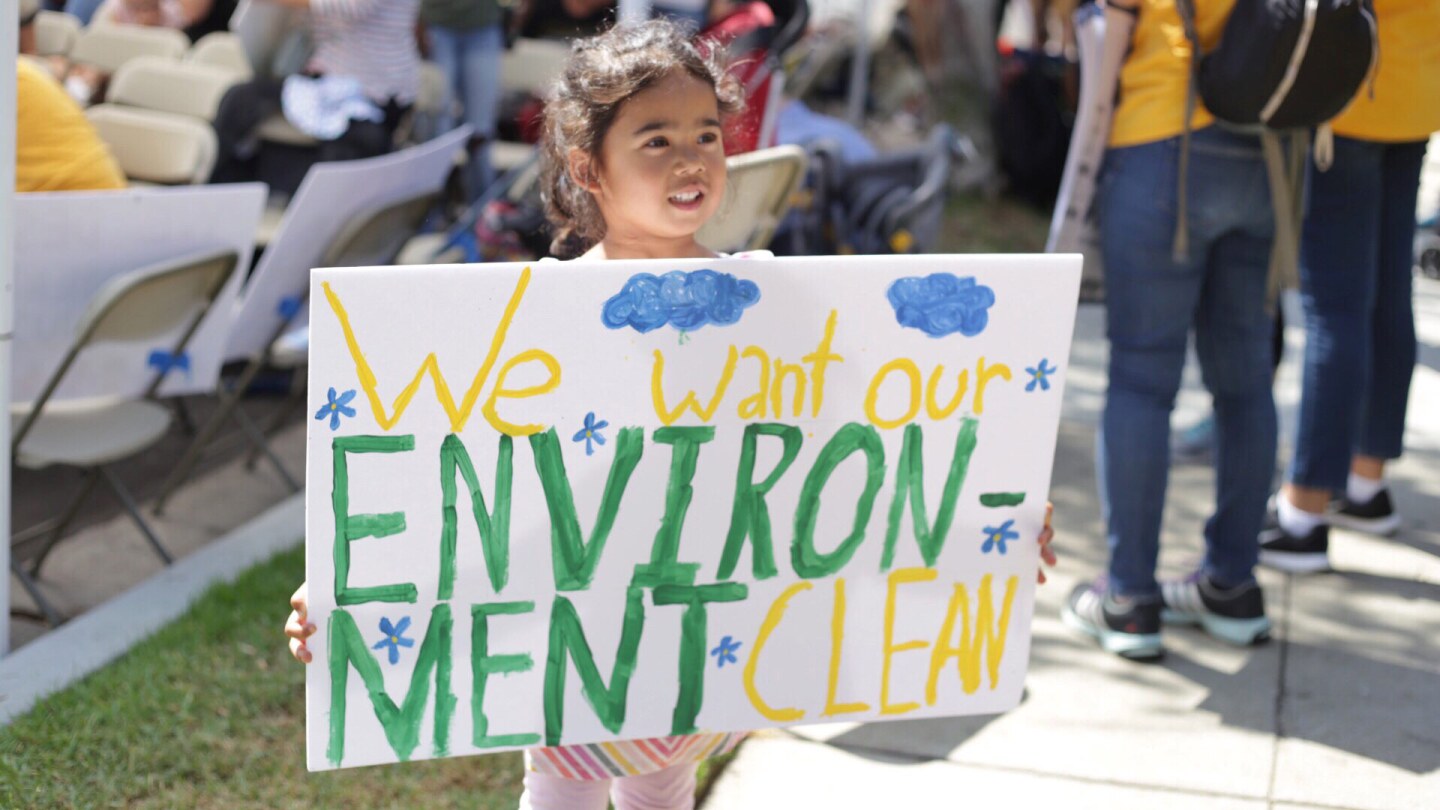 A small child holds a sign up at a protest. It reads "We want our ENVIRONMENT CLEAN."